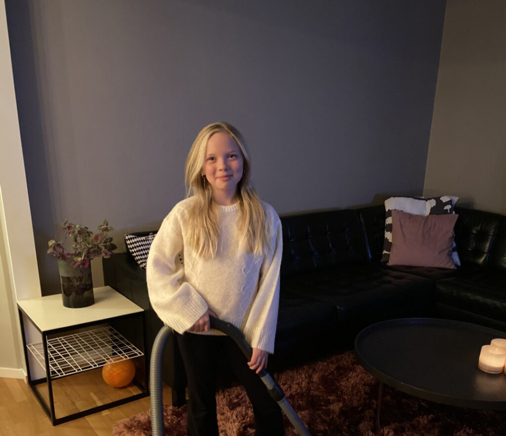 Carmen på Grevhagsskolan håller i en dammsugare. I bakgrunden skymtar ett vardagsrum med en svart soffa.