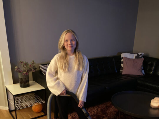 Carmen at Grevhagsskolan is holding a vacuum cleaner. In the background, you can see a living room with a black sofa.