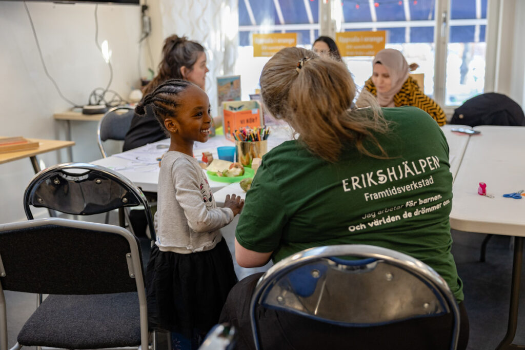 Sarah, som är verksamhetssamordnare, sitter vid ett bord och pratar med ett barn. Bordet är fyllt med färgglada pennor och leksaker.