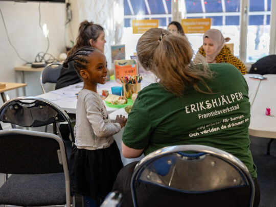 Sarah, who is the program coordinator, is sitting at a table talking to a child. The table is covered with colorful crayons and toys.
