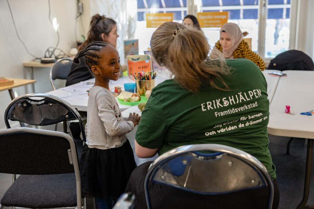 Sarah, who is the program coordinator, is sitting at a table talking to a child. The table is covered with colorful crayons and toys.